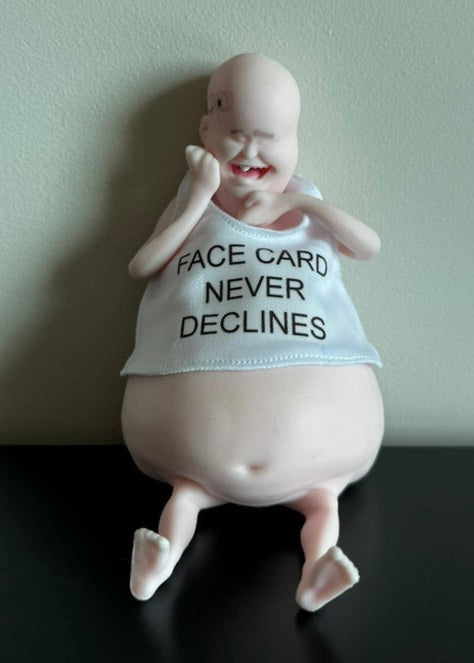 A novelty baby figurine sitting on a black surface against a light green wall. The figurine has a bald head, chubby belly, and is wearing a light blue tank top with the text 'FACE CARD NEVER DECLINES' in black.