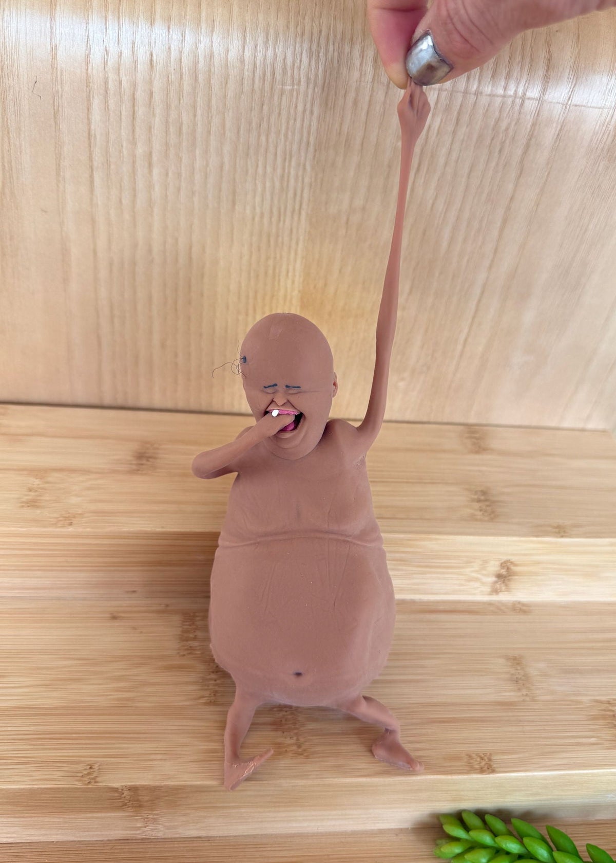 A small flesh-toned novelty figurine or toy with a bald head, wrinkled face, and long thin limbs, standing on a bamboo surface with a hand pulling one of its long arms upward.