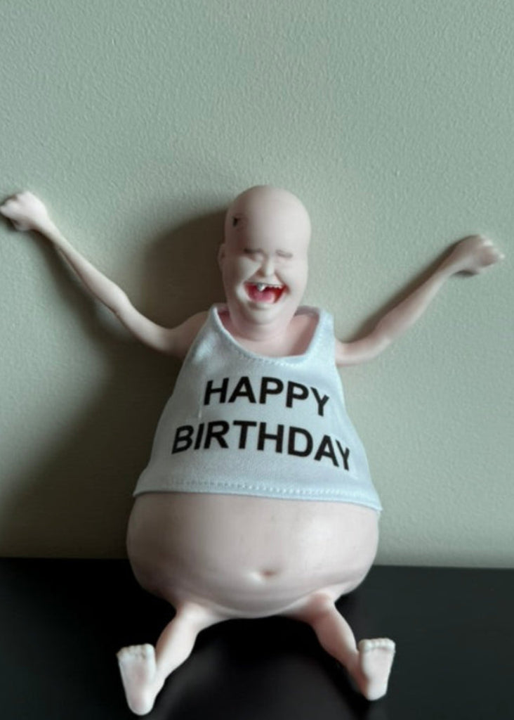 A novelty birthday figurine of a bald, smiling man with outstretched arms, wearing a white tank top that says 'HAPPY BIRTHDAY' in black text, sitting on a black surface against a light green wall.