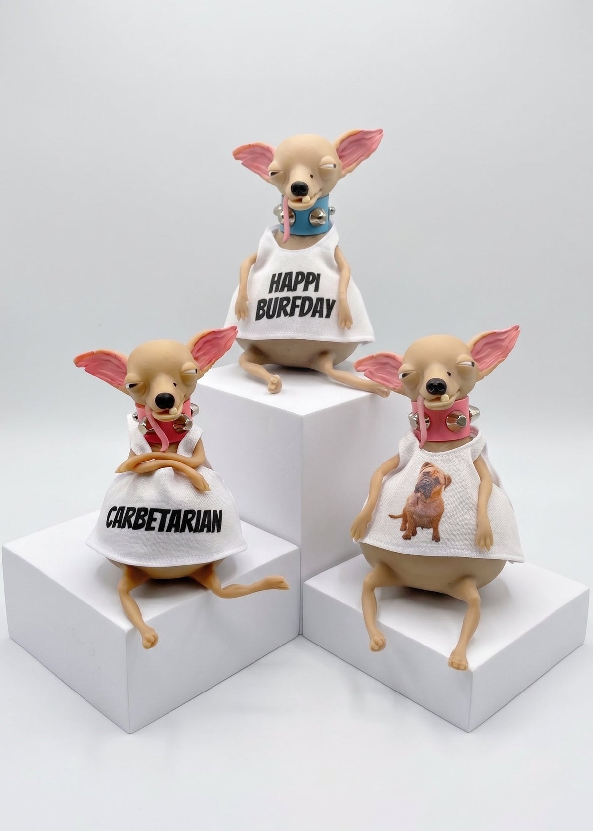 Three small dog figurines posed on white display blocks against a light gray studio background. Each figurine wears a white dress with different printed designs: one says 'CORBETARIAN,' one says 'HAPPY BIRTHDAY,' and one shows a small dog illustration.