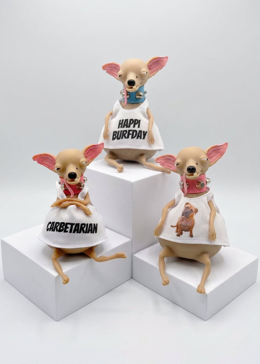 Three small dog figurines posed on white display blocks against a light gray studio background. Each figurine wears a white dress with different printed designs: one says 'CORBETARIAN,' one says 'HAPPY BIRTHDAY,' and one shows a small dog illustration.
