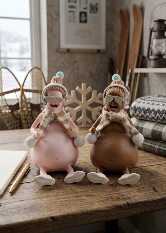 Two whimsical winter-themed figurines sitting on a rustic wooden table. The figurines have round bodies, knit winter hats and scarves, and are styled in a cozy, rustic room setting with snowshoes, a lantern, and plaid blankets in the background.