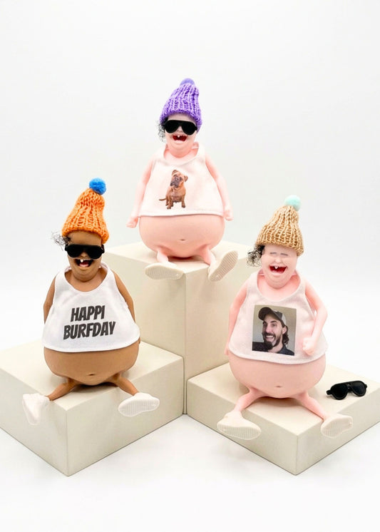 Three novelty figurines of obese cartoon characters wearing knit hats and sunglasses, sitting on white display blocks against a white background. Two figurines wear white tank tops with 'HAPPY BURFDAY' and a photo print, and one wears a pink tank top with a dog print.