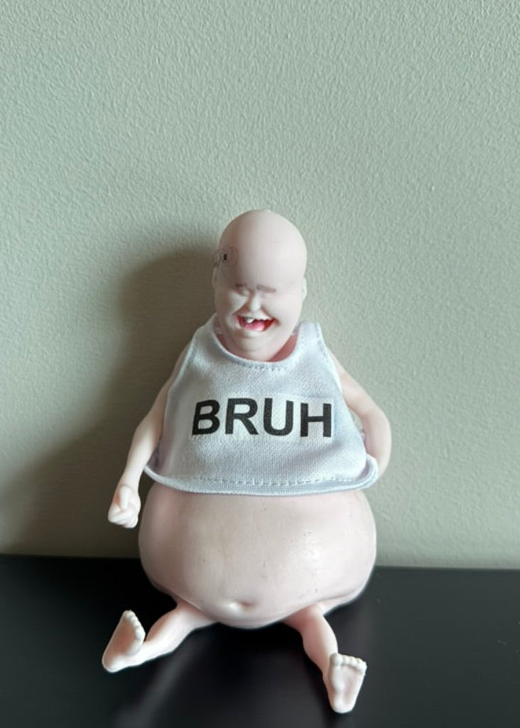 A novelty collectible figurine of a smiling, bald baby-like character with a large belly, wearing a white bib printed with the word 'BRUH' in black. The figurine is sitting on a black surface against a light sage green wall.